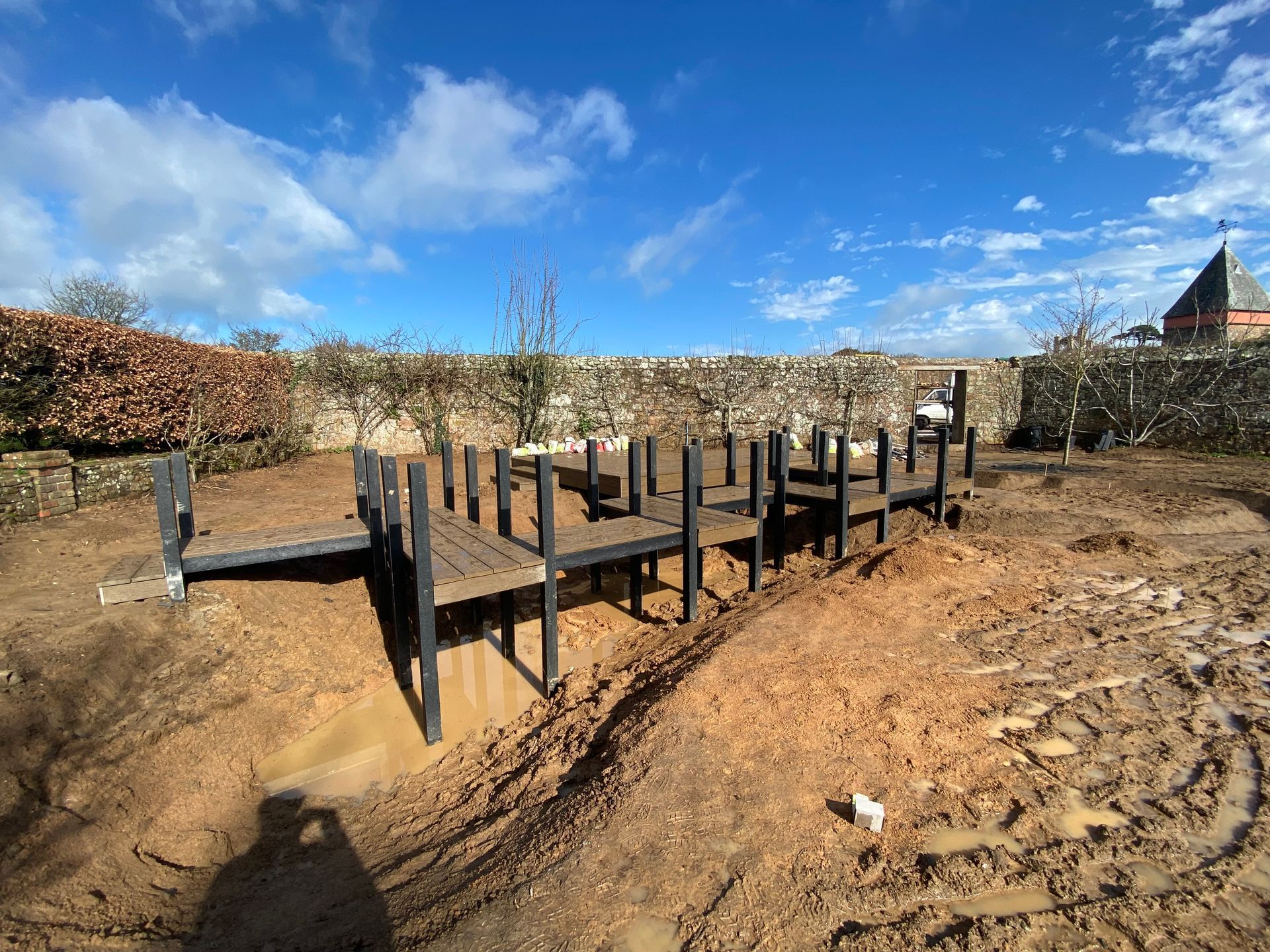 Wooden playground structure in a muddy yard, set against a stone wall and blue sky.