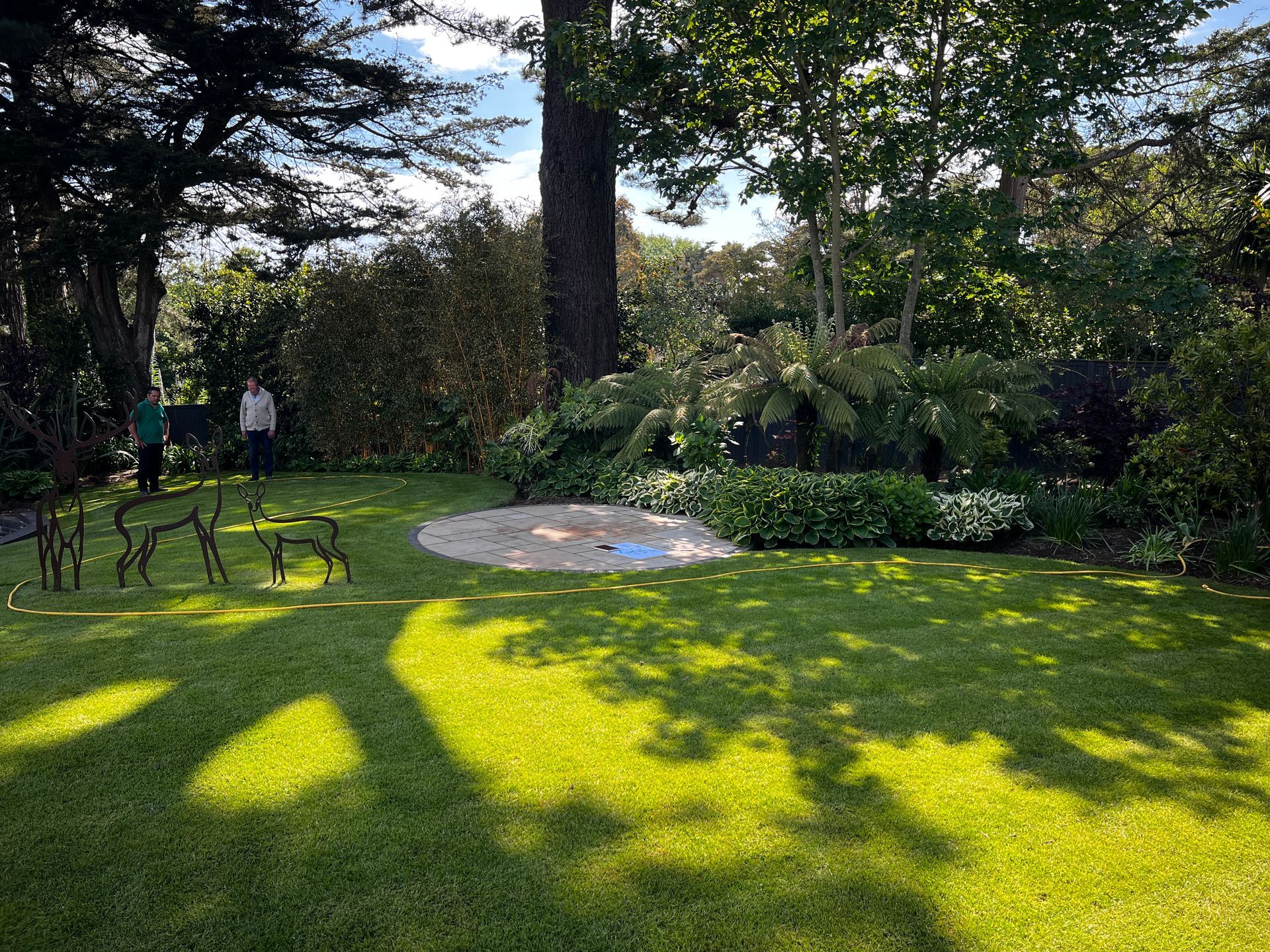 Lush green yard with a fire pit, trees, and two people standing in the background. Deer sculptures in foreground.