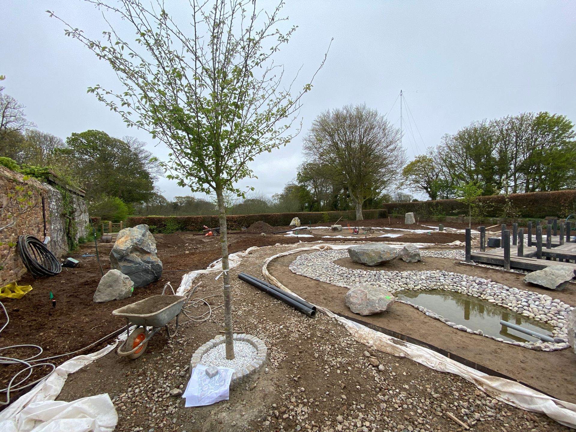 Garden under construction with tree, rocks, gravel, small pond, and wheelbarrow under cloudy sky.