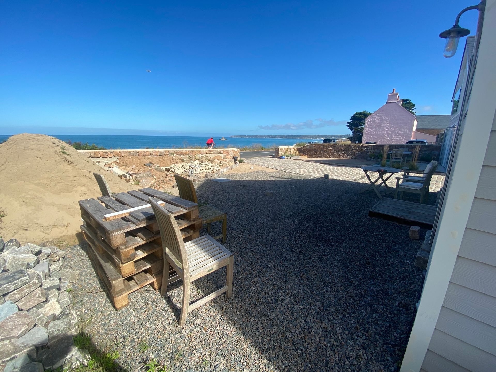 Outdoor scene with sea view, gravel patio, wood pallets, chair, pink building, and clear blue sky.