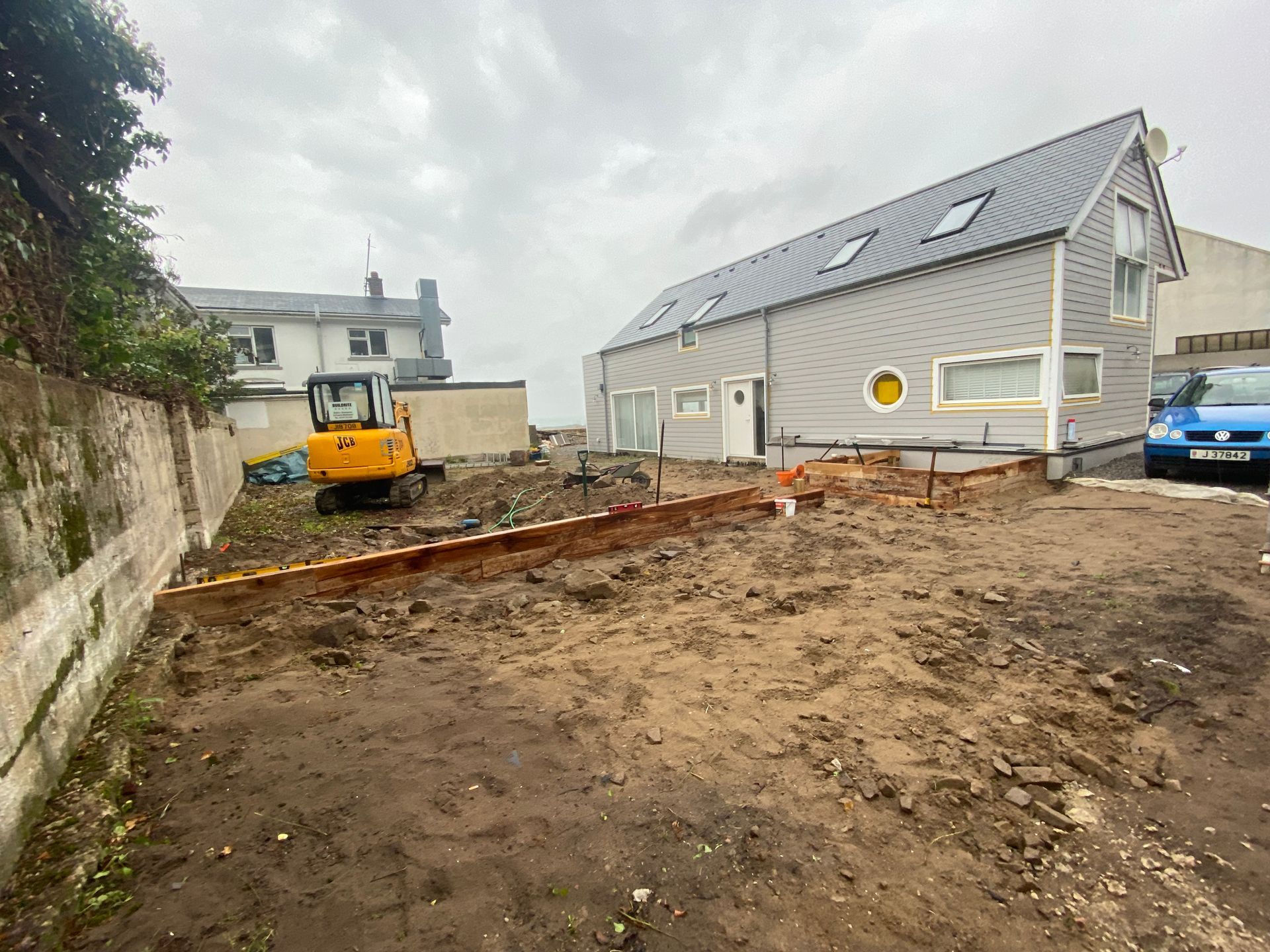 Construction site with a small excavator, next to a grey building with a sloped roof.
