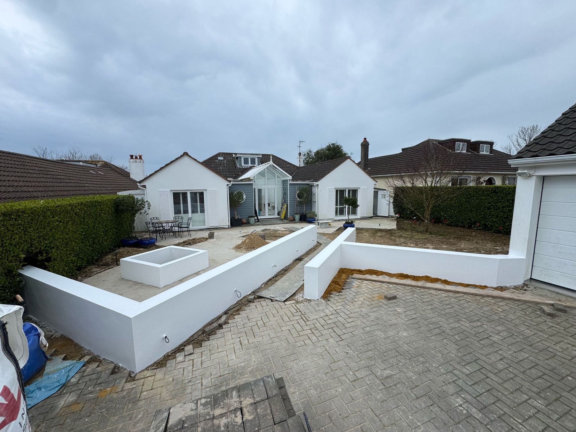 White bungalow with a newly built white wall and a paved driveway under a cloudy sky.