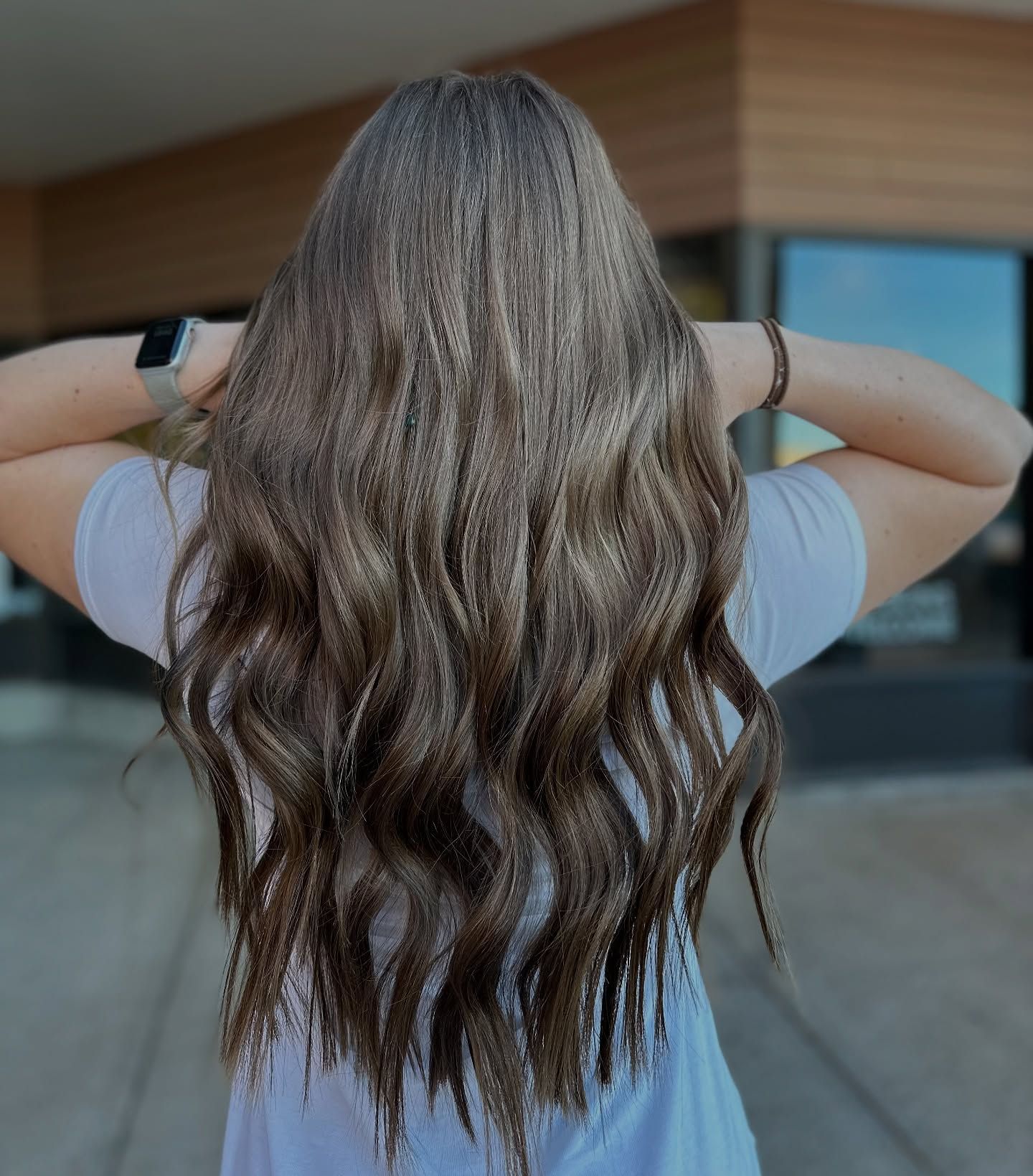 A woman with long brown hair is standing with her hands behind her head.