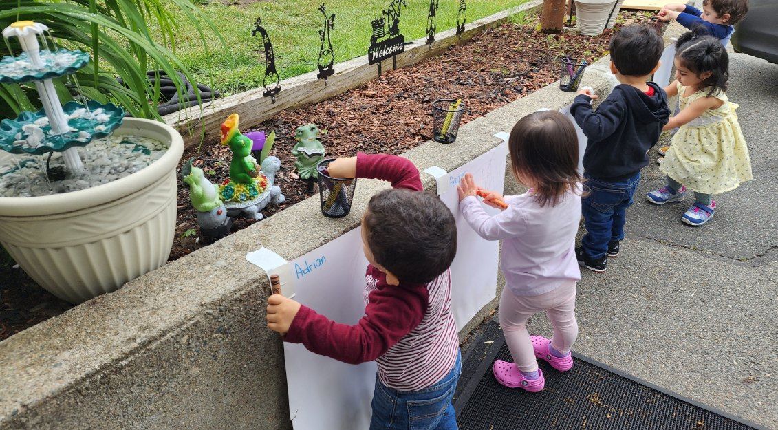 Children painting on a large sheet of paper outdoors near a garden.