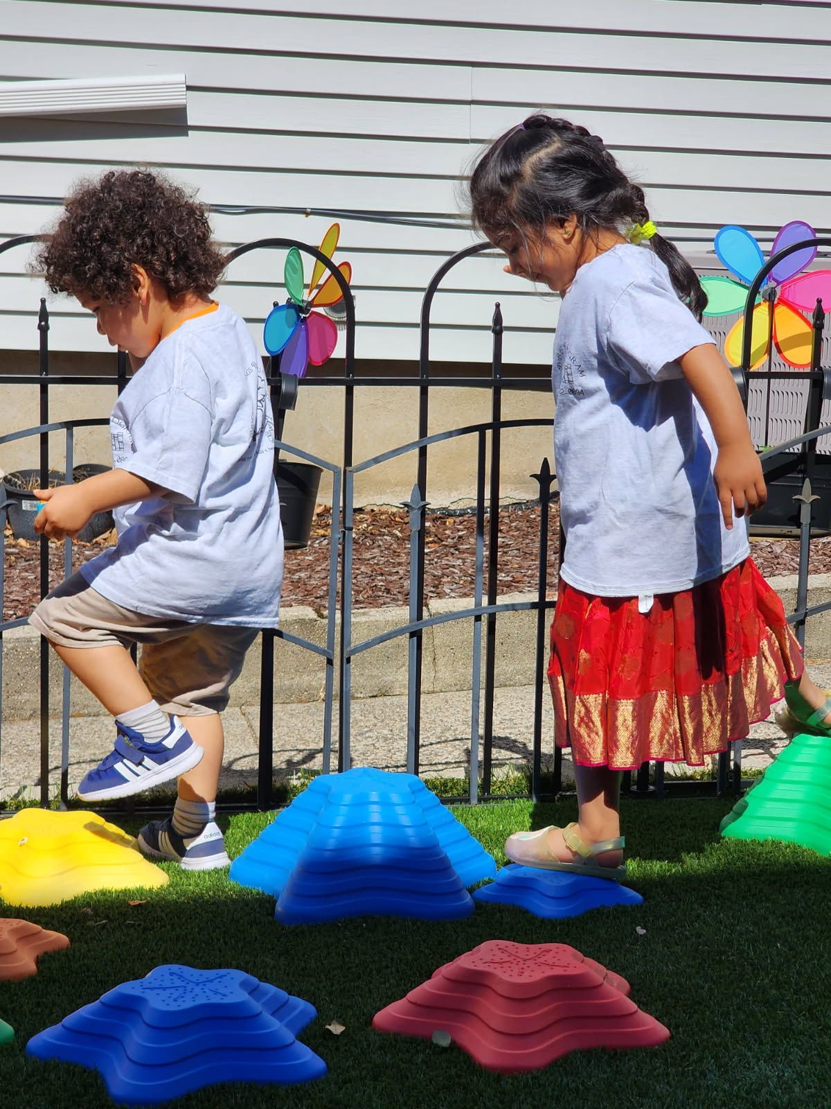 Two children balance on colorful stepping stones in a yard, near a fence with pinwheel decorations.