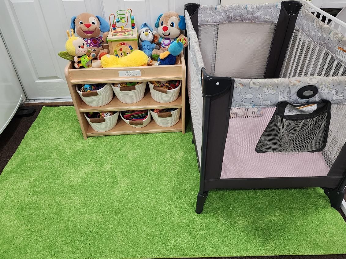 A playpen and toy shelf on a green rug. Toys are on top shelf and in white baskets below.