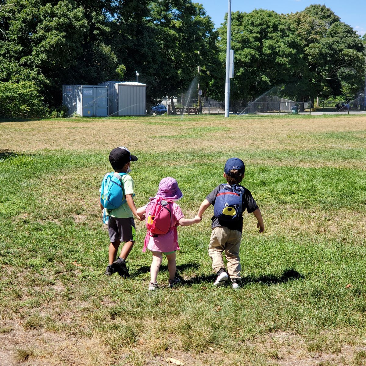 Three children holding hands walking on grass towards a sprinkler, wearing backpacks.
