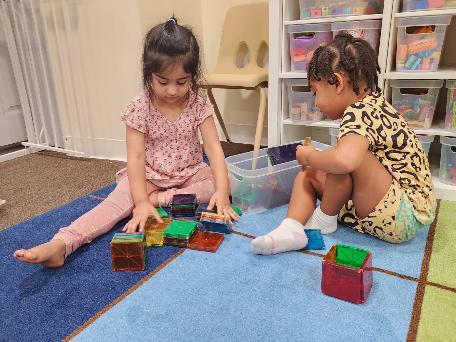 Teacher tossing a ball to three smiling children sitting on a blue patterned rug in a classroom.