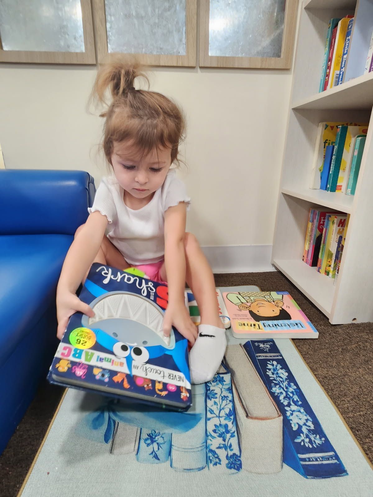 Young child in white top and pink shorts reading a book on the floor. Bookshelf in background.