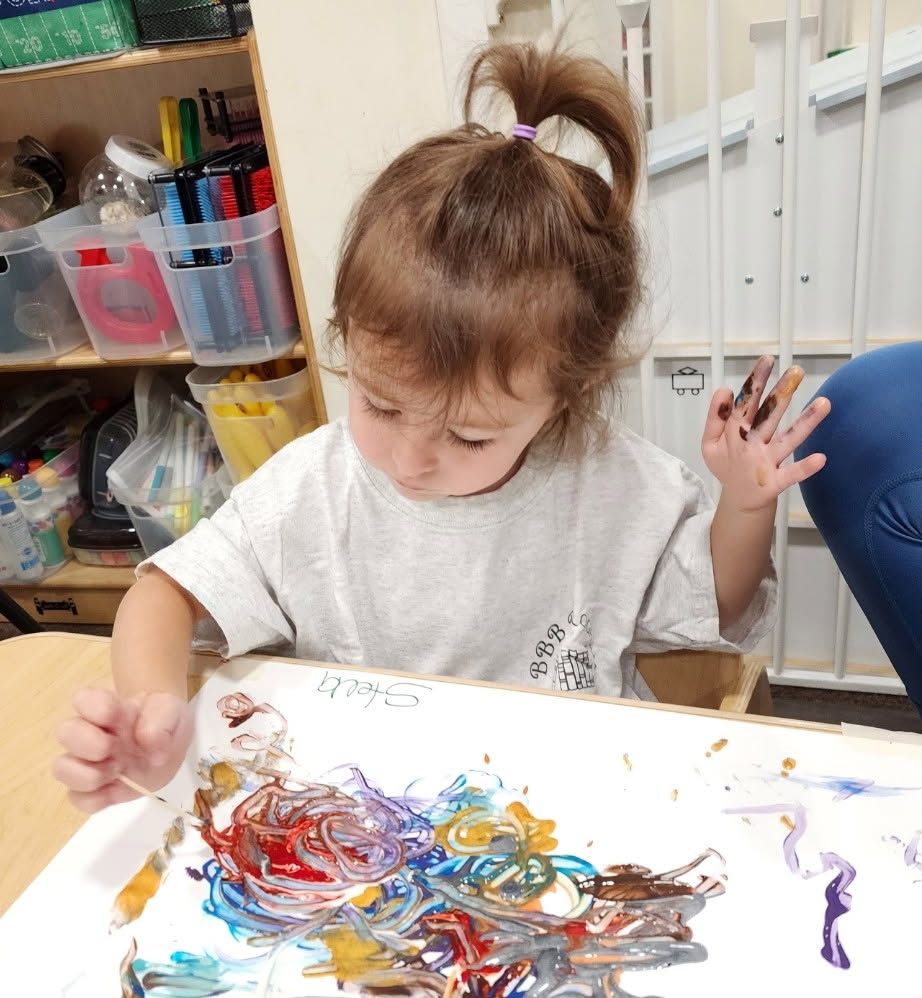 Child painting with hands, wearing gray shirt, with paint-covered hands and artwork on a table.