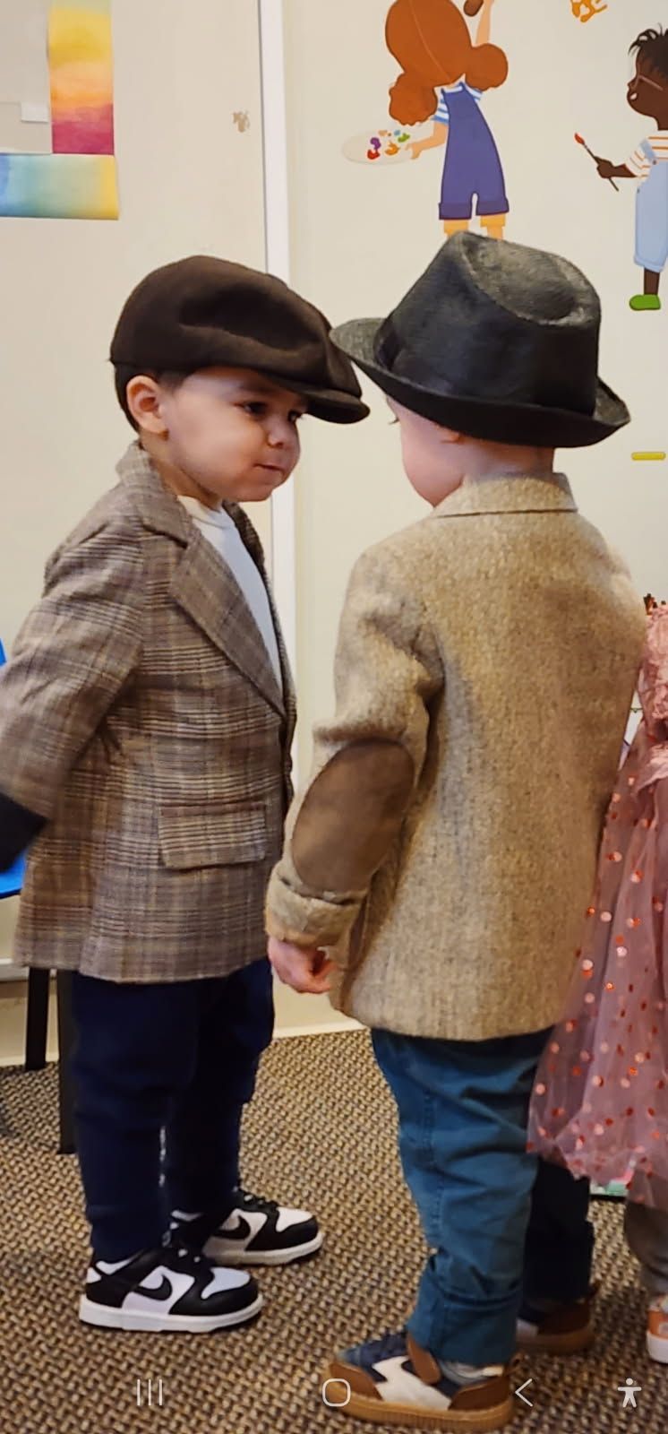 Two children in vintage-style jackets and hats face each other, indoors, near a wall with art.