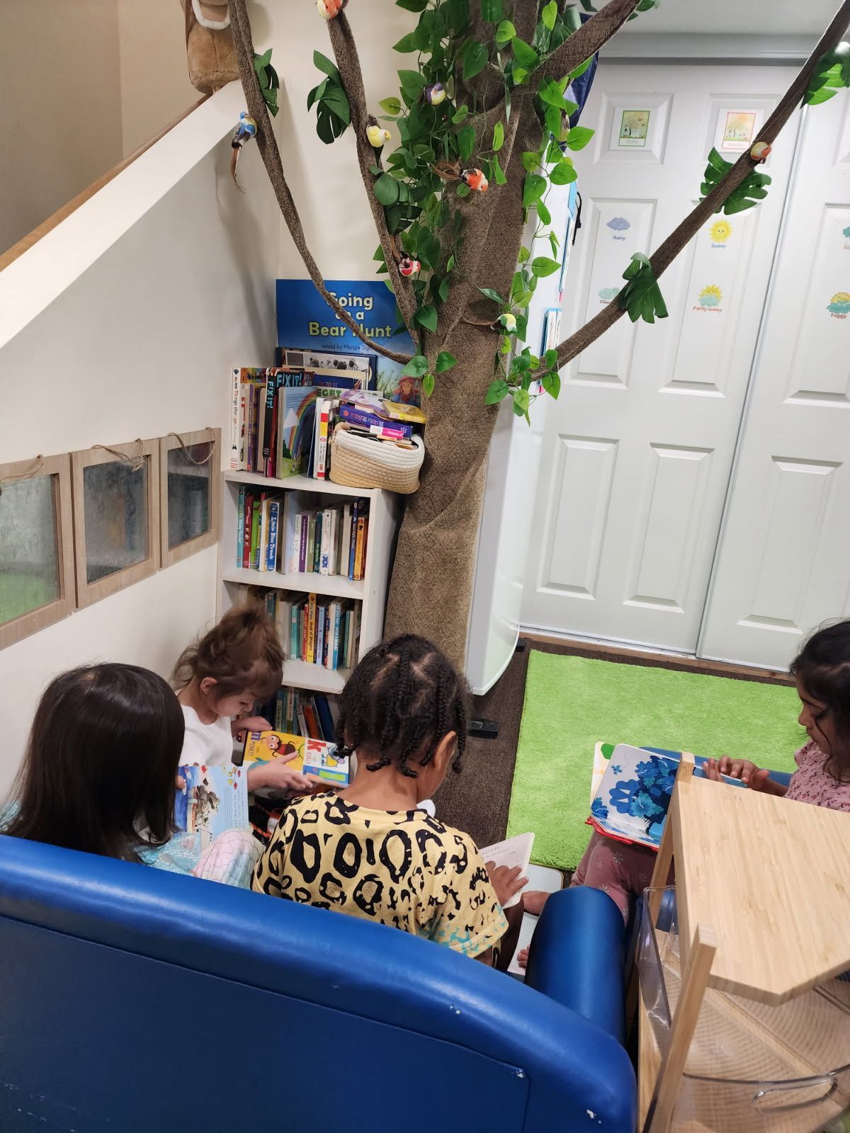 Children reading in a cozy corner with a tree trunk, books, and a blue slide.