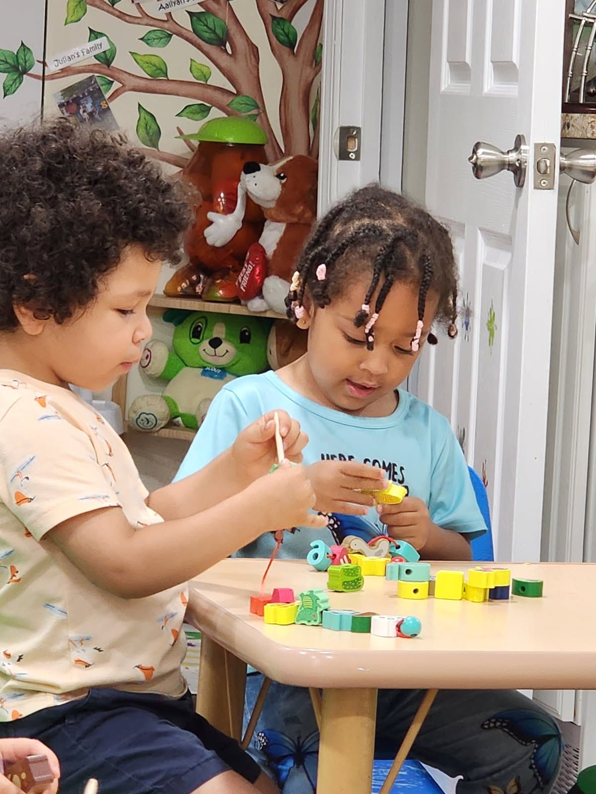 Two children playing with colorful blocks at a table in a room with a tree mural.
