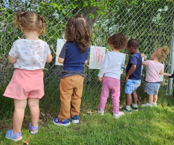 Teacher assisting four children drawing at a table, with colorful artwork on the wall.