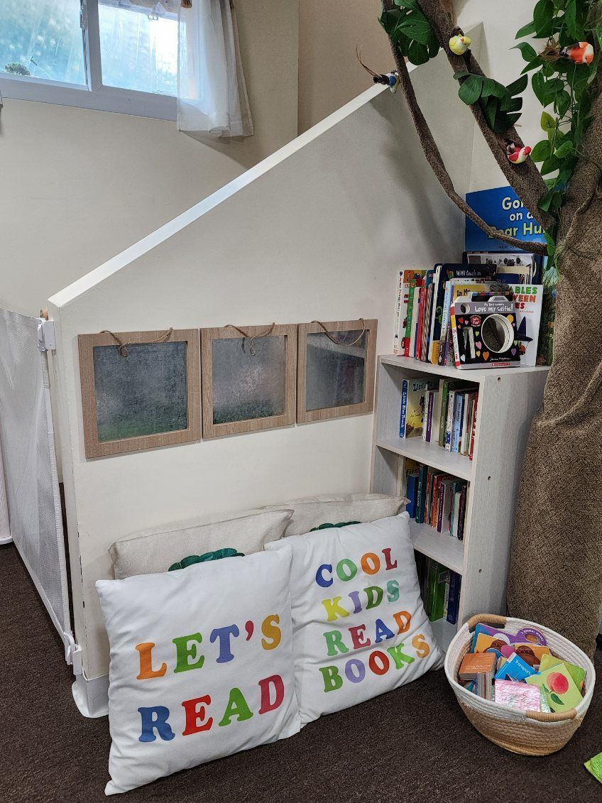 Cozy reading nook with a house-shaped structure, pillows, and a bookshelf. Includes tree decor and a basket of toys.