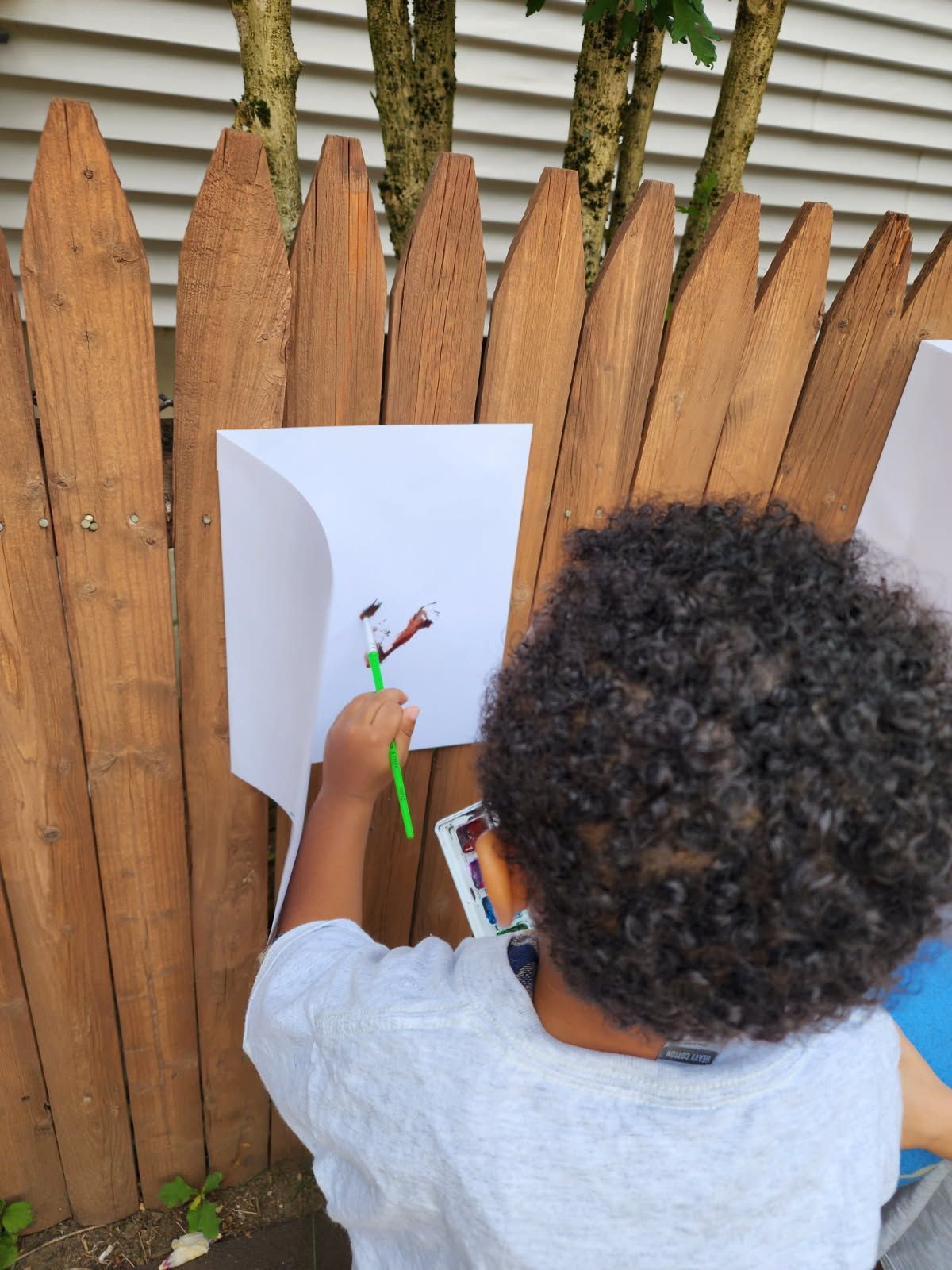 Child with curly hair painting a picture attached to a wooden fence with a green paintbrush.