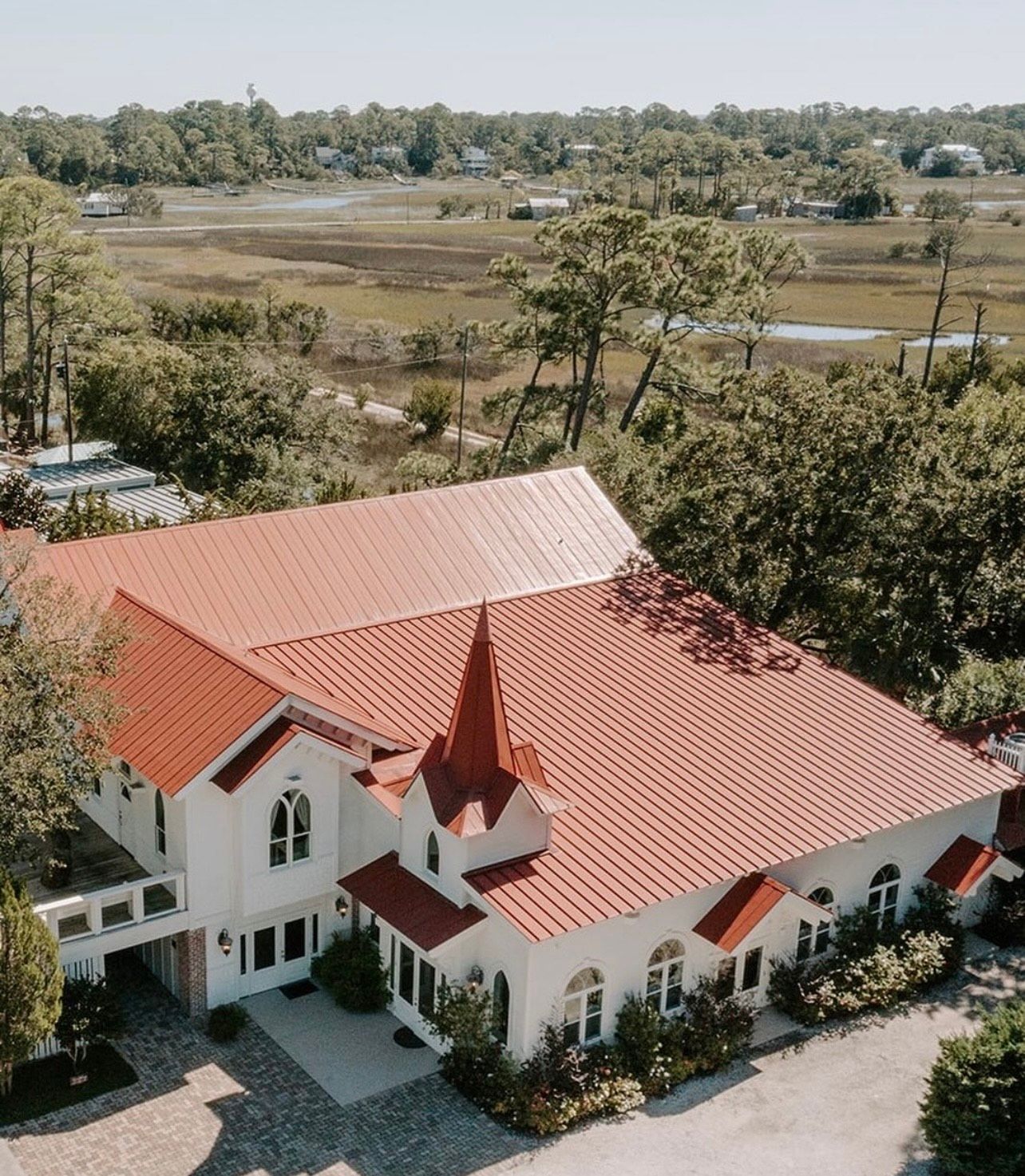 White building with red roof, steeple, and arched windows, surrounded by trees and marsh.