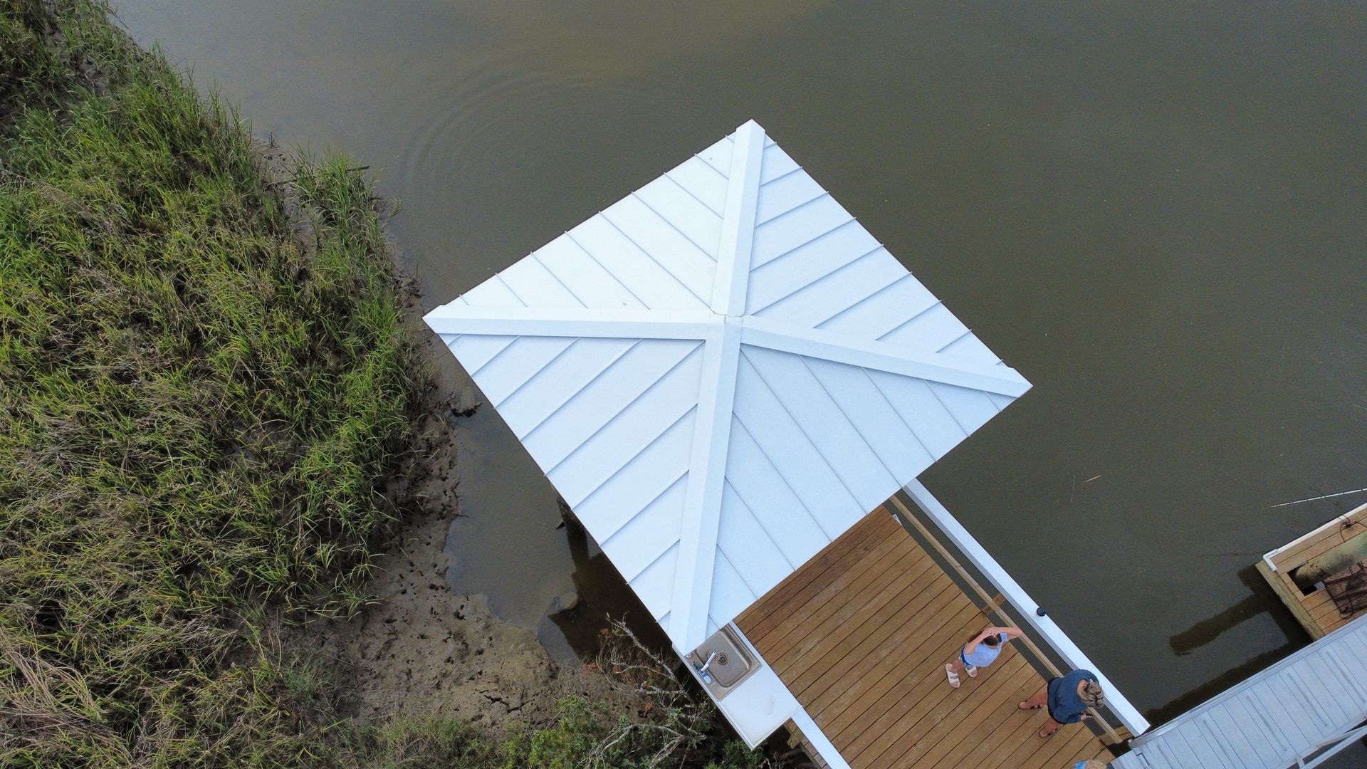 Overhead view of a dock with a white, peaked roof. A person relaxes on the wooden deck beside the water.