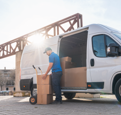 A delivery worker in a blue uniform loads a stack of cardboard boxes onto a hand truck next to a white cargo van.