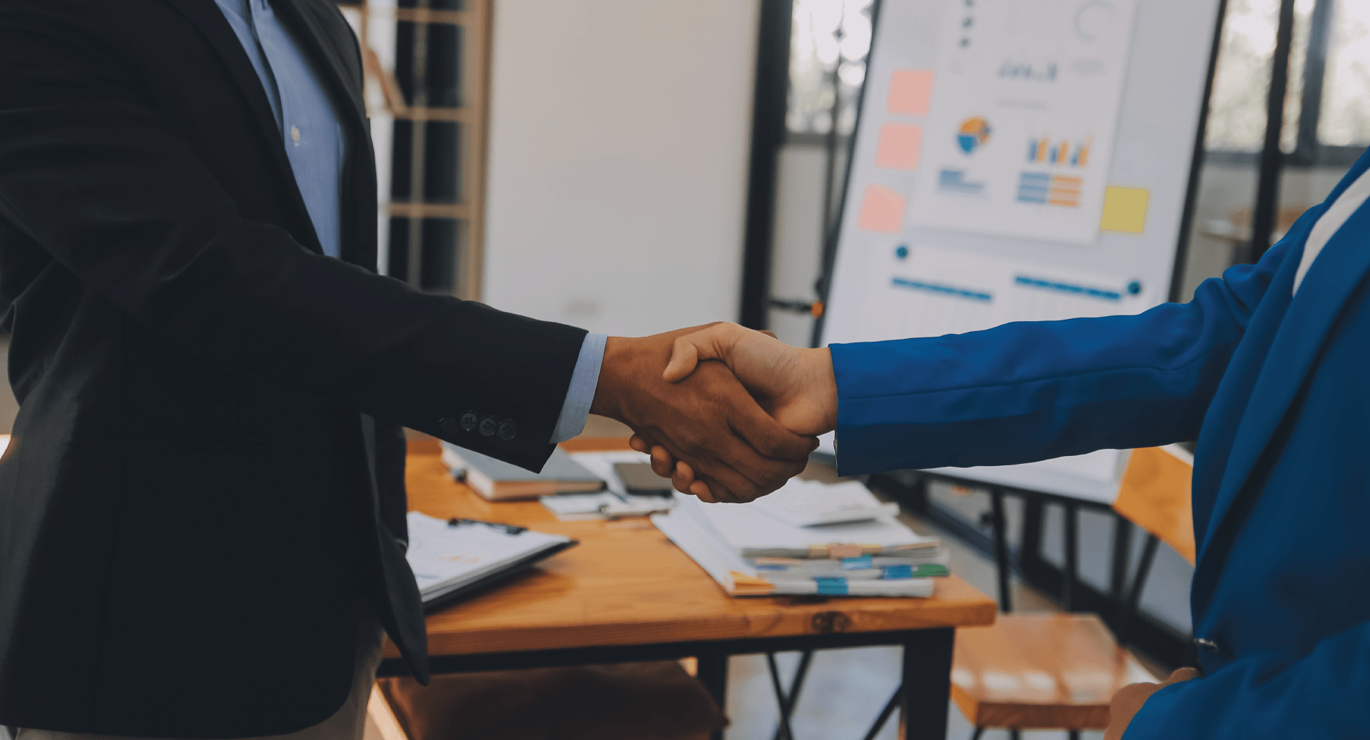 Two business professionals in professional attire shake hands over an office desk with papers and a whiteboard nearby.