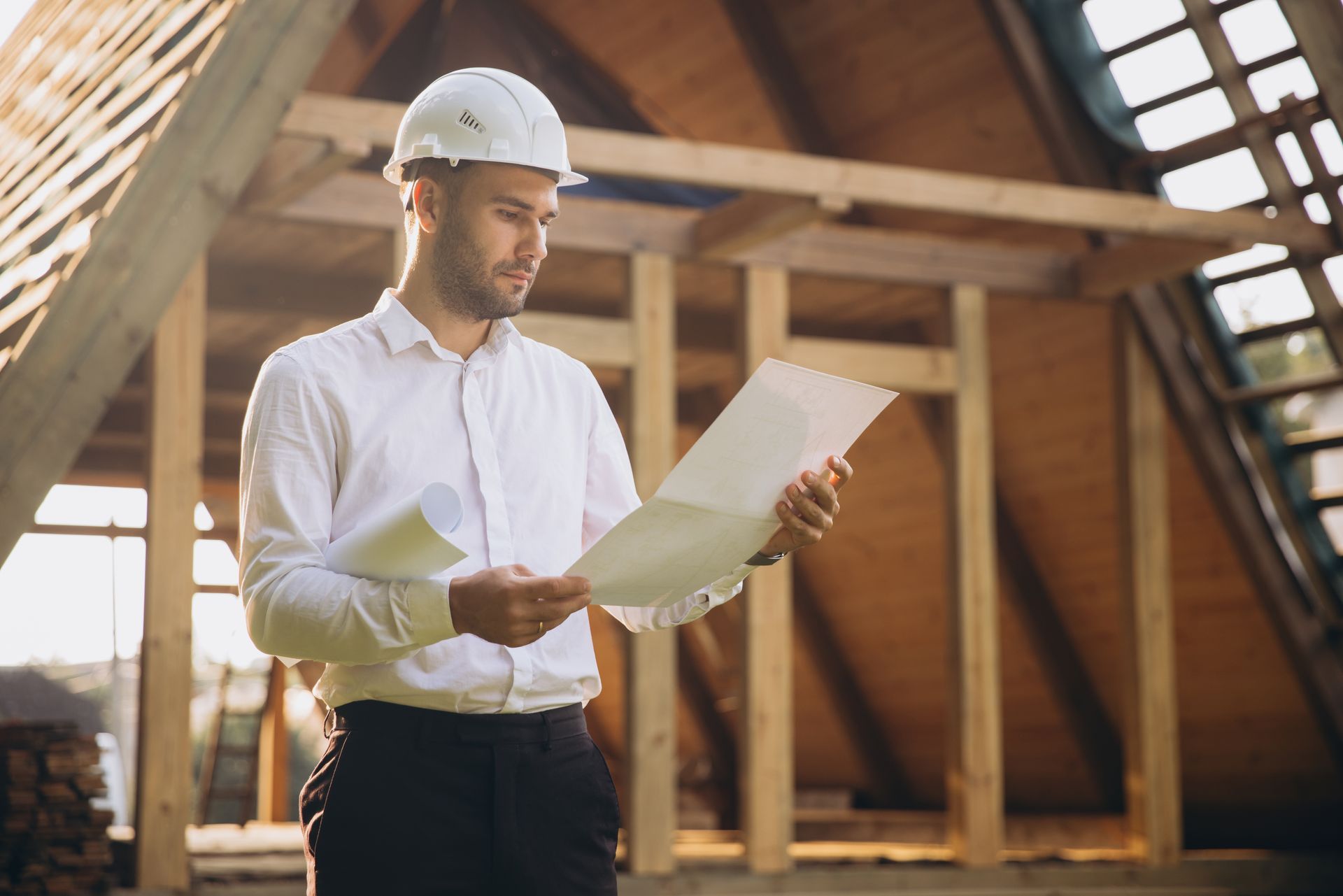 A person in a hard hat and button-down shirt stands inside a wooden building frame, examining architectural blueprints.