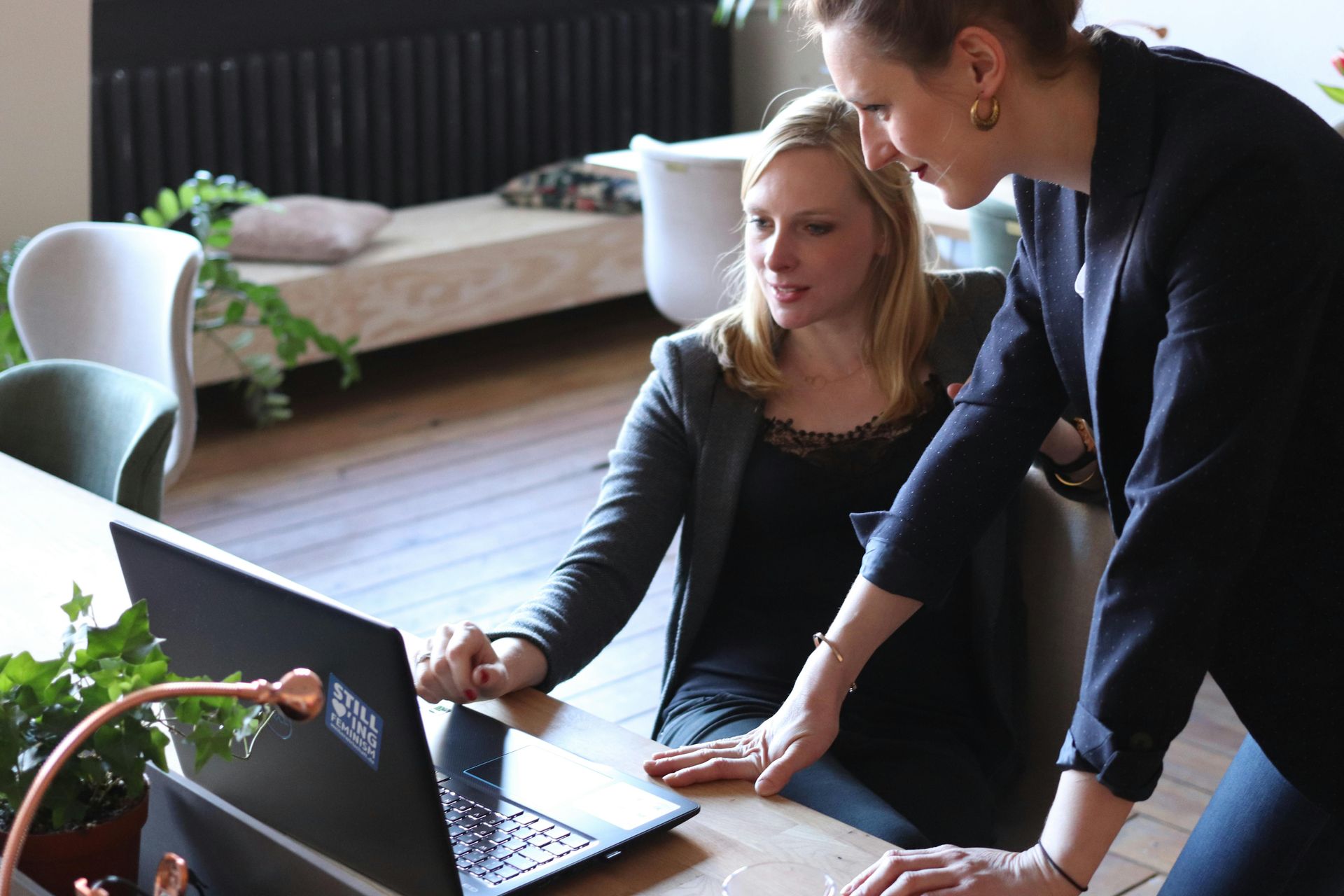 Two professional women collaborate while looking at a laptop in a modern, plant-filled office space.