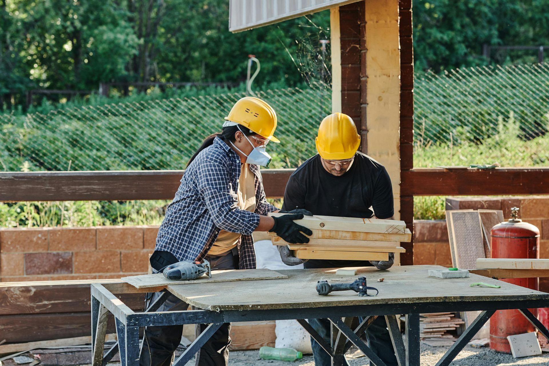 Two people wearing yellow hard hats and safety gear work together with wooden planks on a table at an outdoor construction site.
