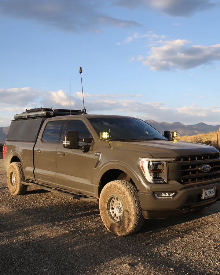 A tan Ford F-150 off-road truck with a bed cap and equipment, parked on a gravel path against a desert mountain backdrop.