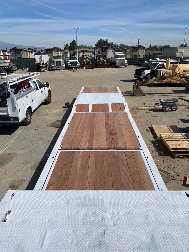 A high-angle view of a flatbed trailer featuring wooden decking, parked in a sunny industrial construction yard.