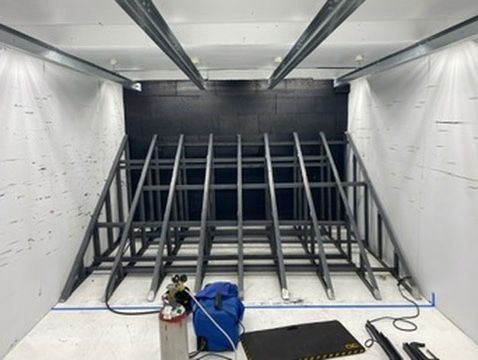 A metal framework structure sits inside a room with white walls, a black back wall, and equipment on the floor.