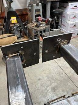 Two steel rectangular tubes with flat metal mounting plates are positioned face-to-face in a workshop setting.