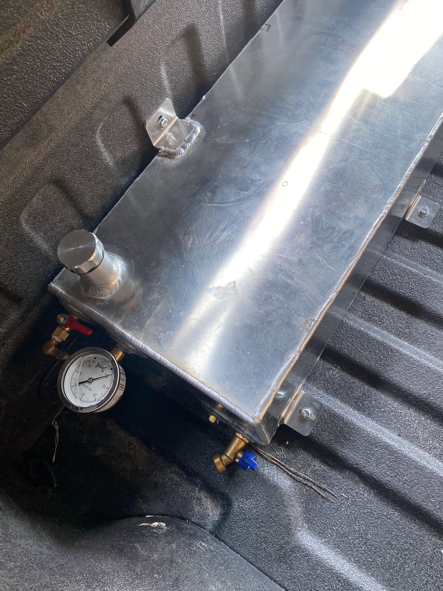 A rectangular aluminum tank with a pressure gauge and two valves, secured inside the corrugated bed of a truck.