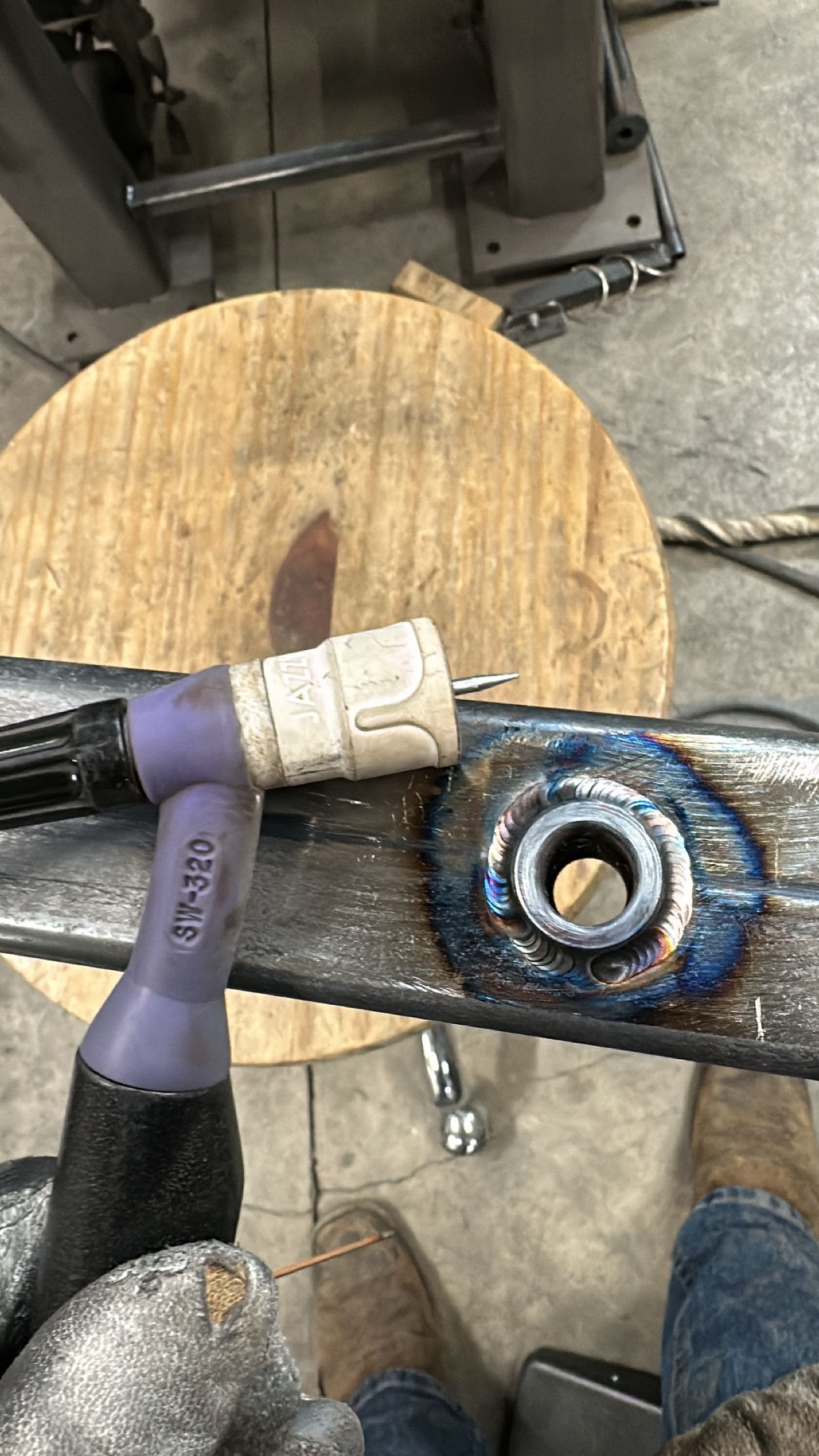 A person holding a purple TIG welding torch next to a metal piece with a circular welded nut on a wooden workbench.