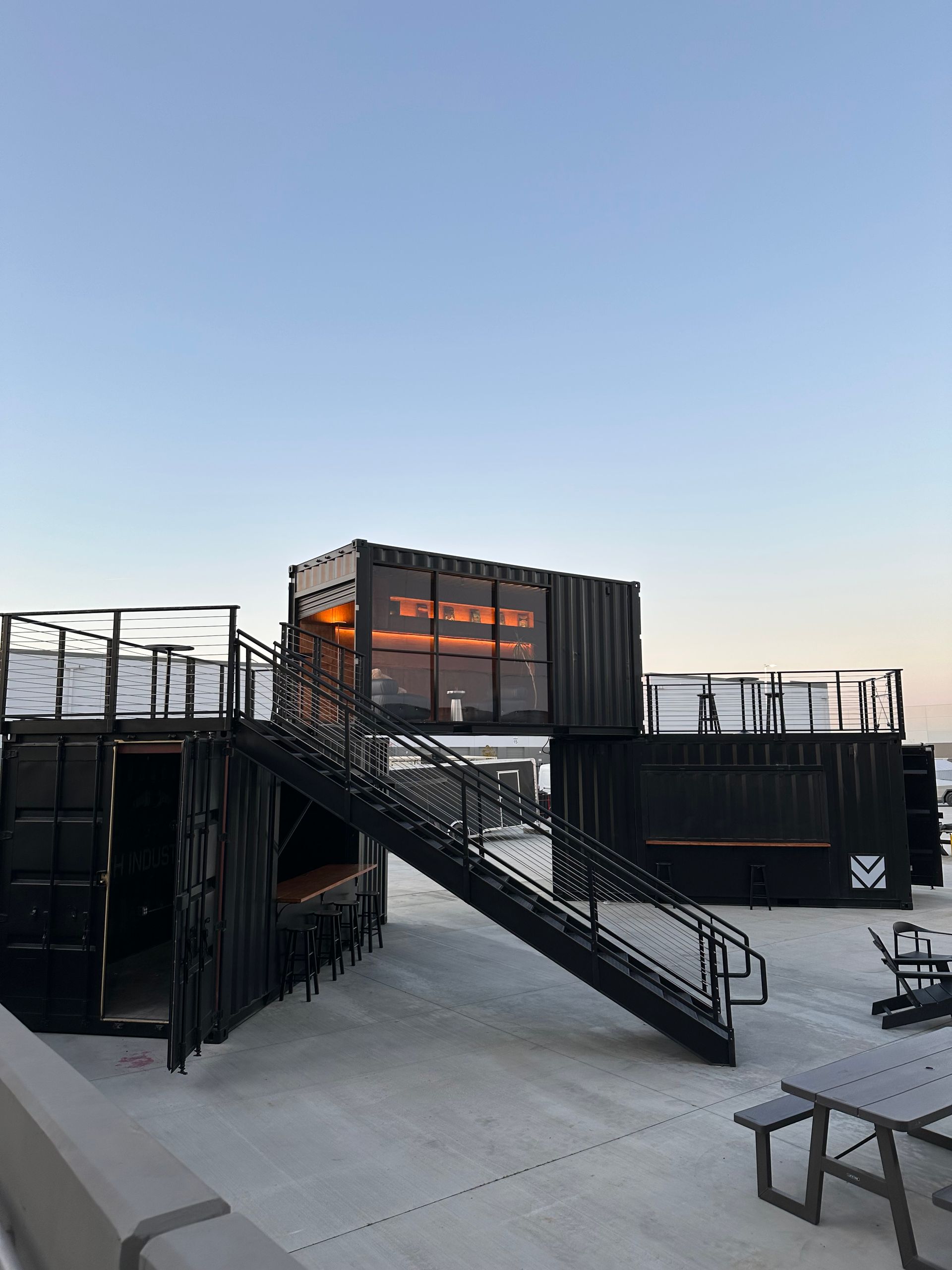 A multi-level outdoor venue made of black shipping containers with staircases, rooftop seating, and warm interior lighting.