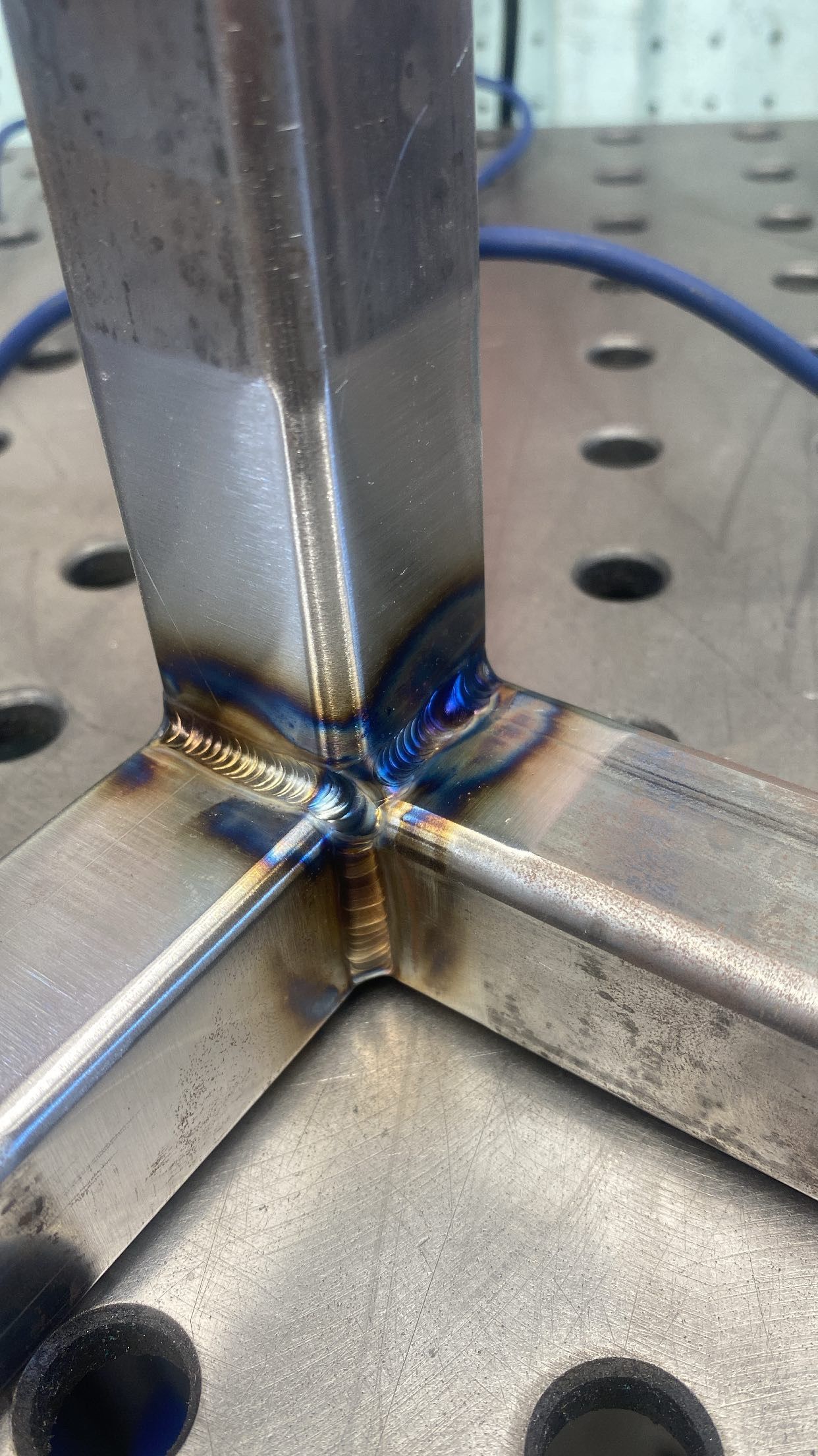 A close-up of a metal square tube welded onto a flat perforated metal table, showing heat-tinted welded joints.