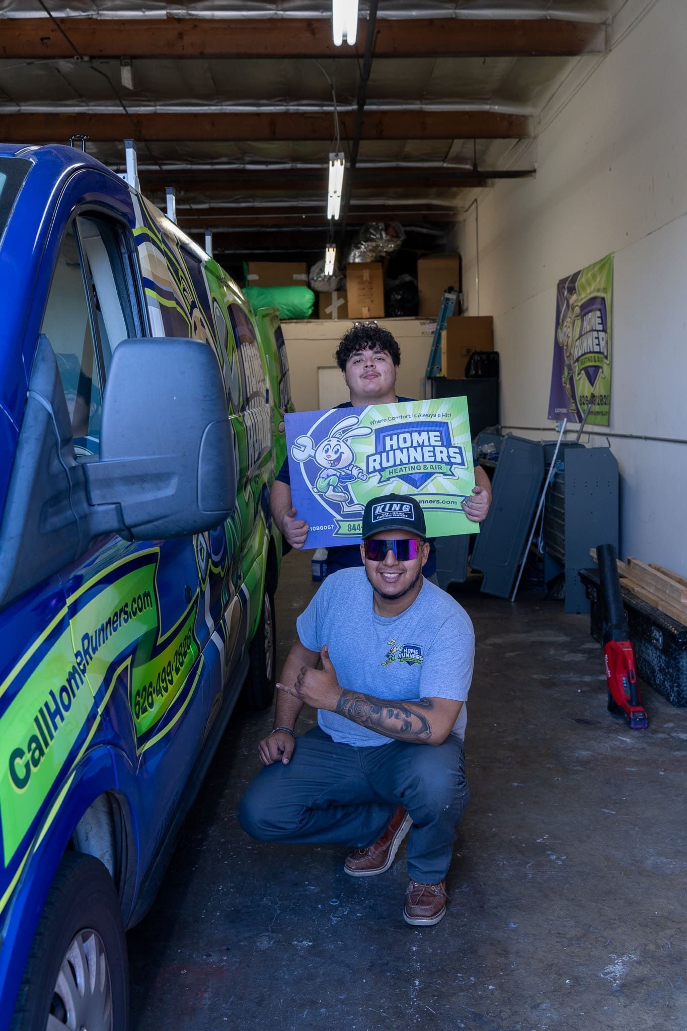 Two people in front of a blue van, one holding a sign. They are in a garage setting.