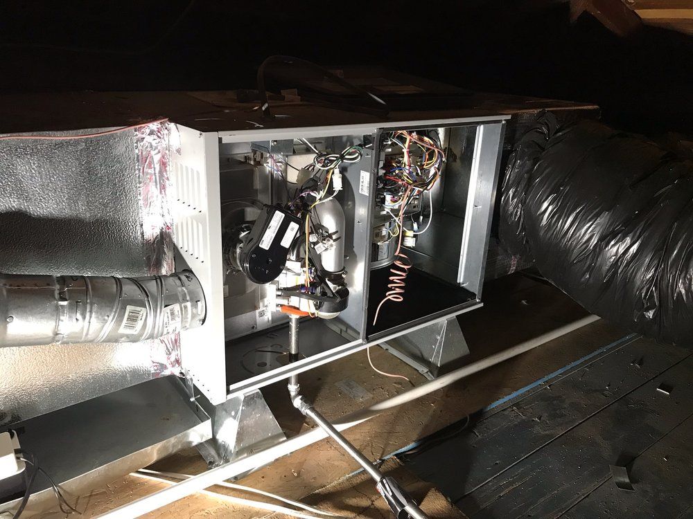 Furnace unit in an attic. Metal box with exposed components, ductwork, and wiring. Dark setting.