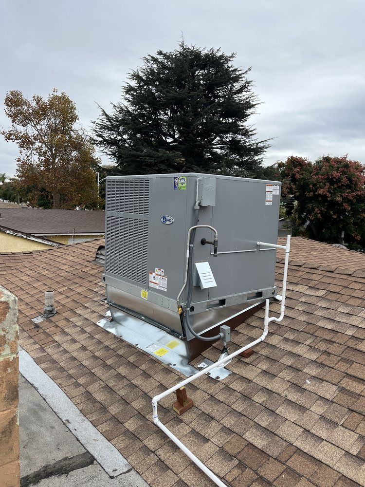 Rooftop HVAC unit on a shingled roof, grey metal box with attached white pipes, overcast sky.