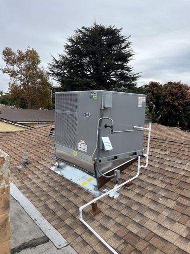 Rooftop HVAC unit on a brown shingle roof; grey unit with white pipes against a cloudy sky.