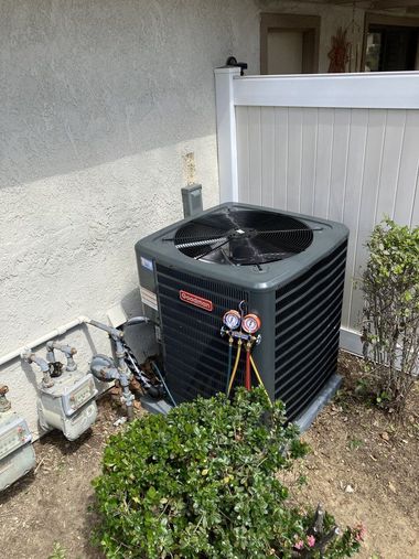 HVAC unit with gauges outdoors near a wall, fence, and shrubs.