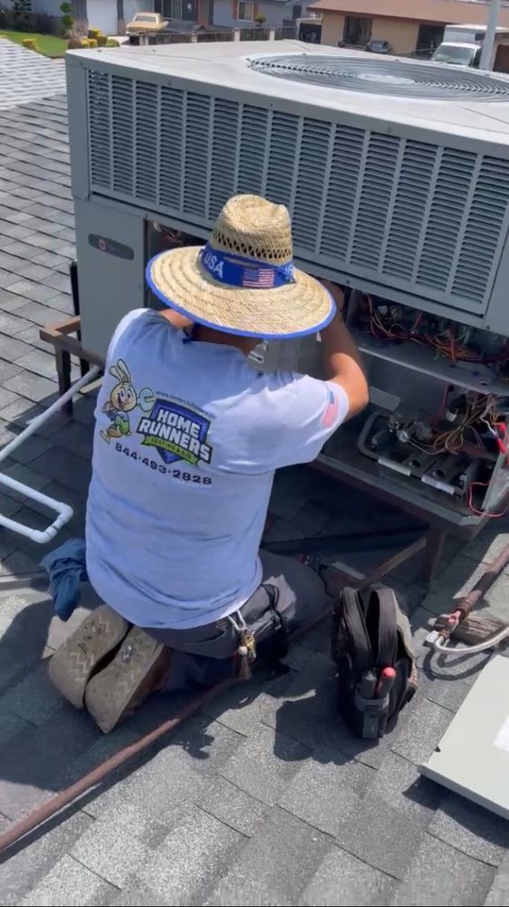 HVAC technician repairing an air conditioning unit on a rooftop. He's wearing a hat, kneeling, and working on the machinery.