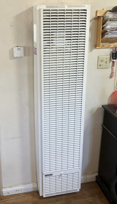 White wall-mounted gas heater on a wall with a thermostat, electrical outlet, and wooden shelf.