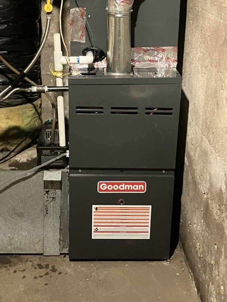 A dark gray Goodman gas furnace in a basement. White pipes and a silver chimney are attached.