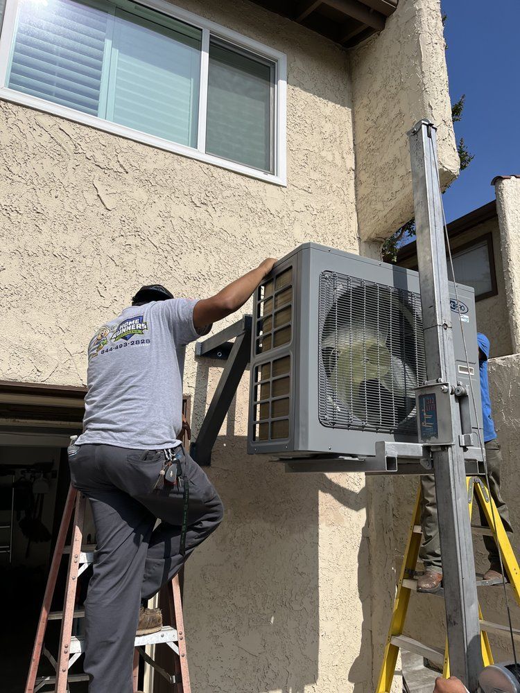 Workers installing a gray air conditioning unit on a building exterior. One worker on a ladder.