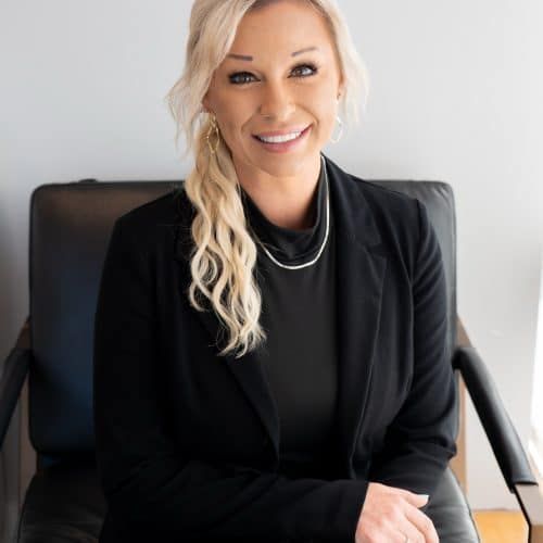 Smiling person in black blazer seated in a chair against a light wall