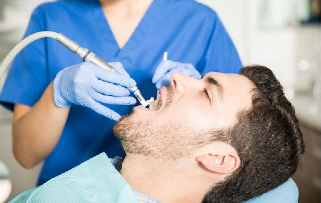 Dentist using a drill on a patient reclining in a chair, wearing blue gloves and scrubs.