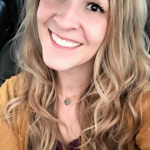 Smiling woman with wavy blonde hair wearing a necklace and tan top, posing for a selfie