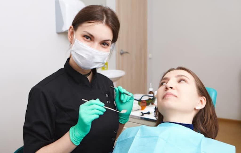Dentist wearing mask and green gloves examines a patient in a dental chair with tools in hand.