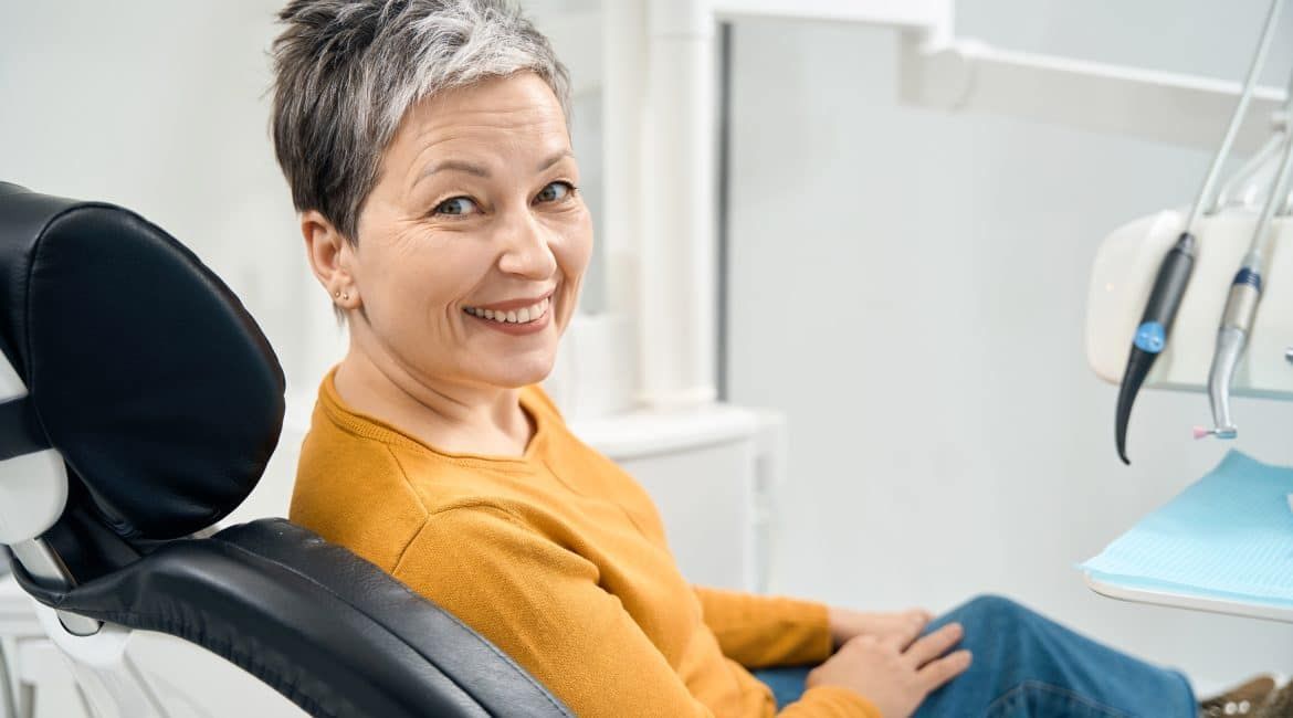 Smiling woman in a dental chair, wearing a mustard sweater, in a bright dental clinic