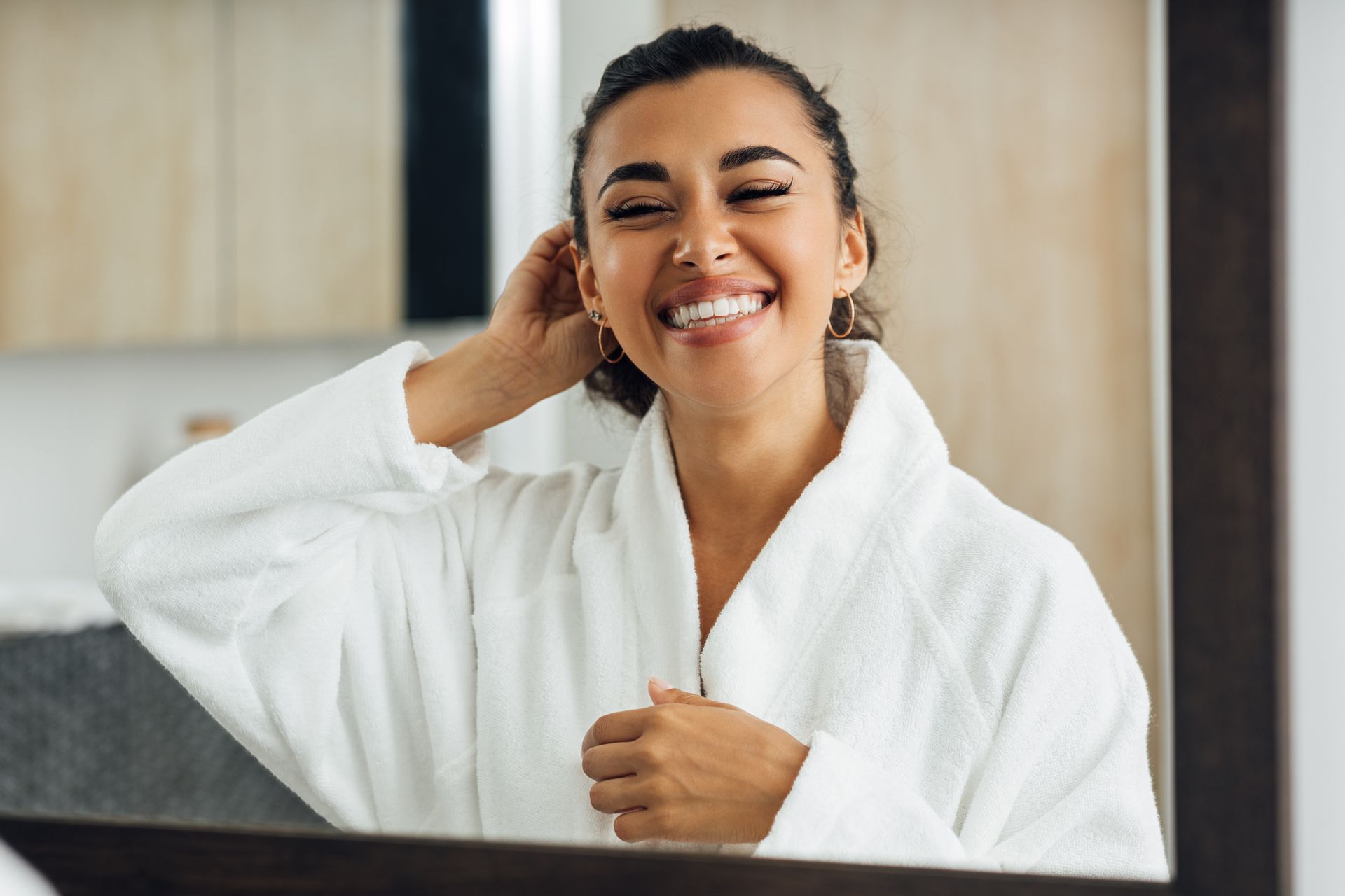 Smiling person in a white bathrobe adjusting hair in a bathroom mirror